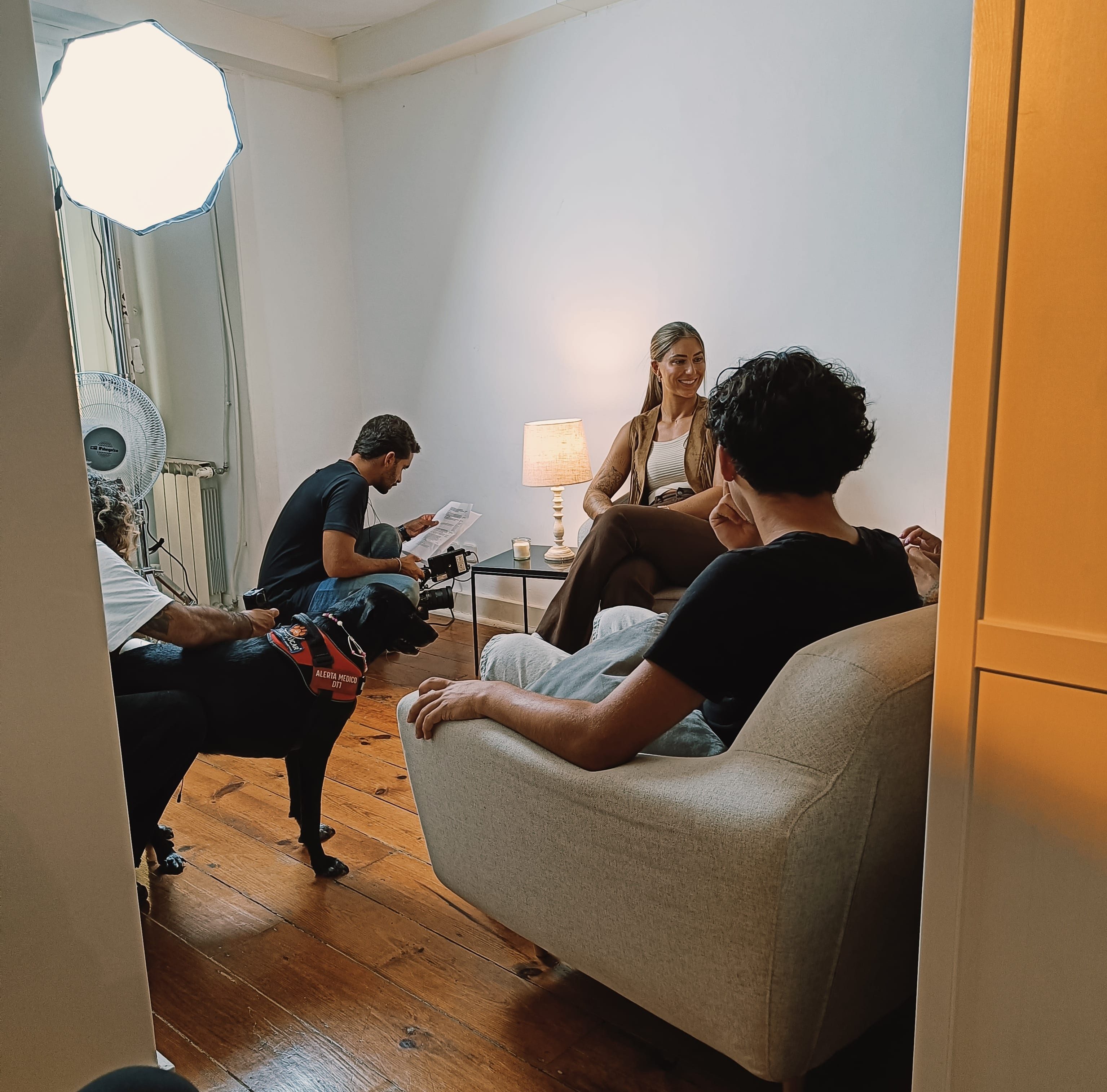 sala onde foi feita a gravação da campanha Focinhos de Açúcar criada pela MAPFRE seguros. Estão um grupo de pessoas numa sala iluminada, sentadas num sofá e em cadeiras, durante uma conversa informal, com um cão preto ao lado e equipamentos de fotografia no canto. Ambiente acolhedor com piso de madeira e luz natural.