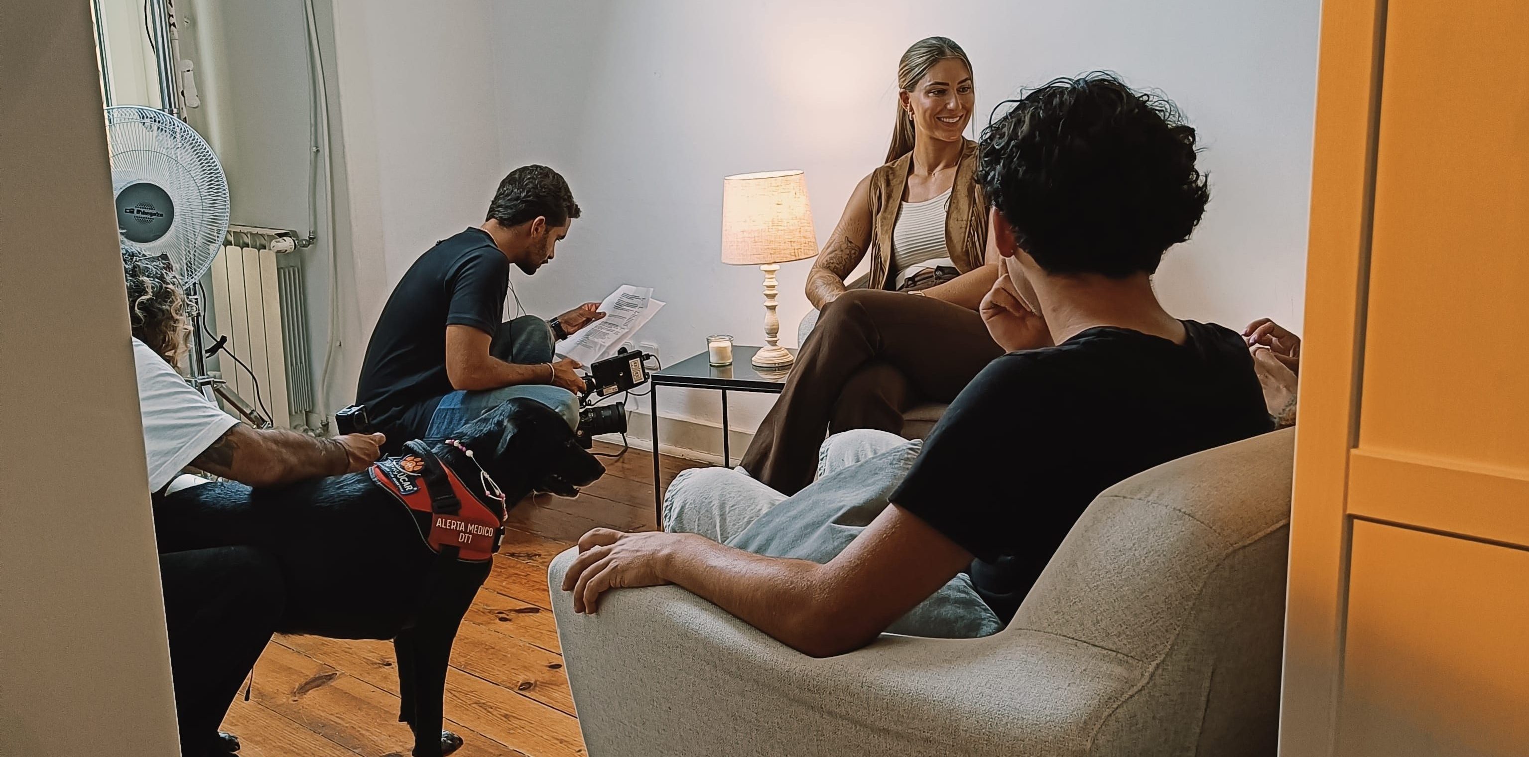 Gravação da campanha Focinho D’Açúcar sala onde foi feita a gravação da campanha Focinhos de Açúcar criada pela MAPFRE seguros. Estão um grupo de pessoas numa sala iluminada, sentadas num sofá e em cadeiras, durante uma conversa informal, com um cão preto ao lado e equipamentos de fotografia no canto. Ambiente acolhedor com piso de madeira e luz natural.
